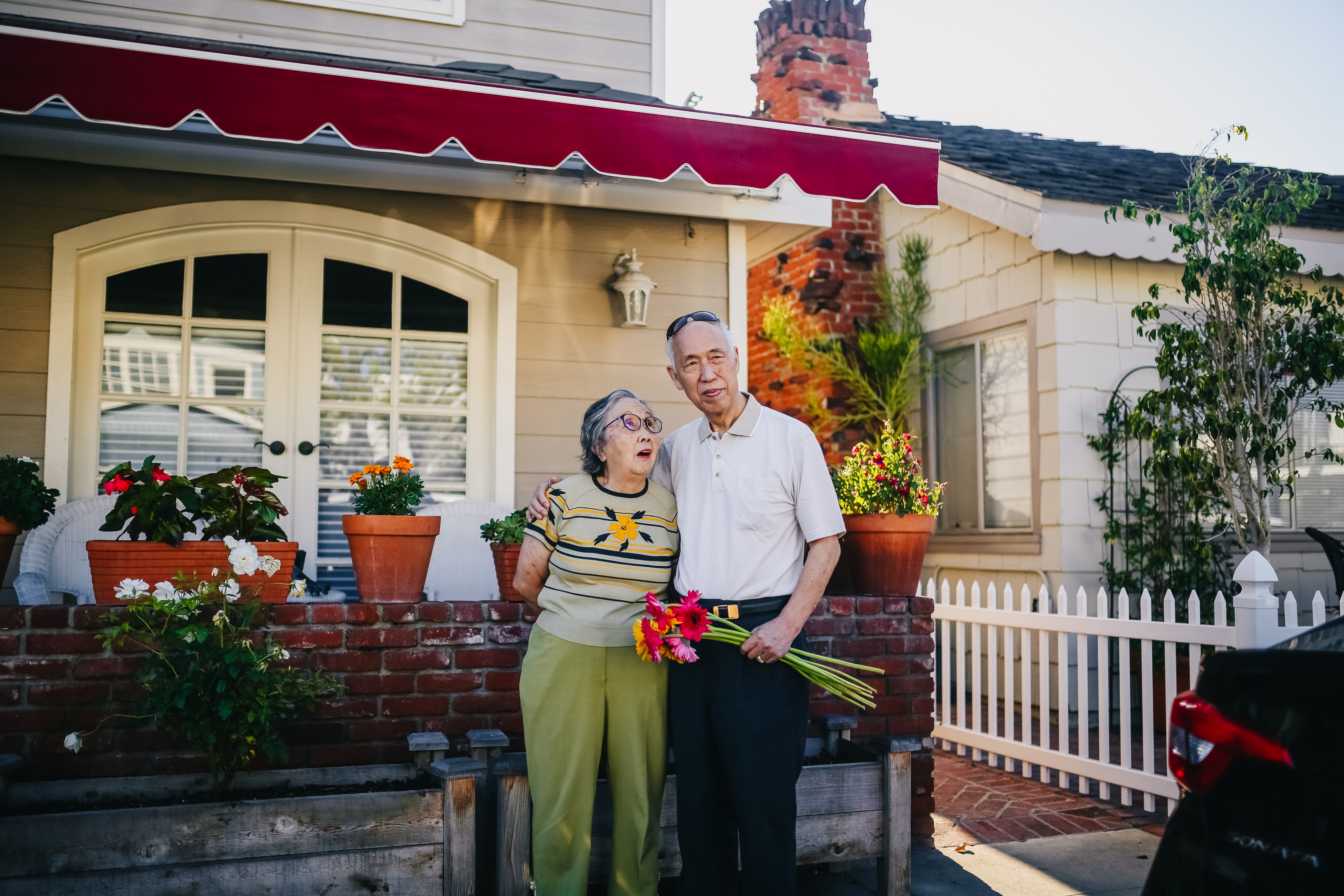 Elderly couple standing outside their home with lots of flowers.