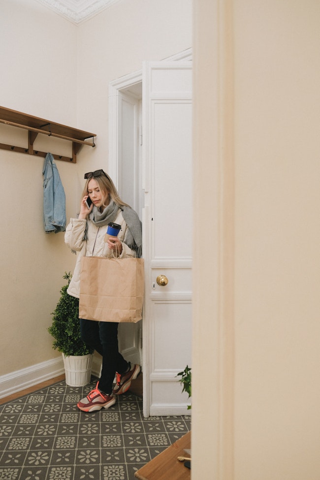 Woman coming into her home or apartment while talking on the phone and carrying a bag.
