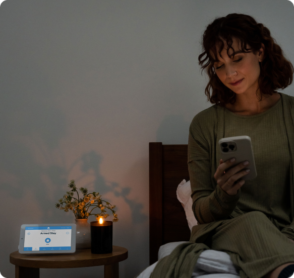 Woman on bed arming her security system with her phone, with the alarm panel next to her.