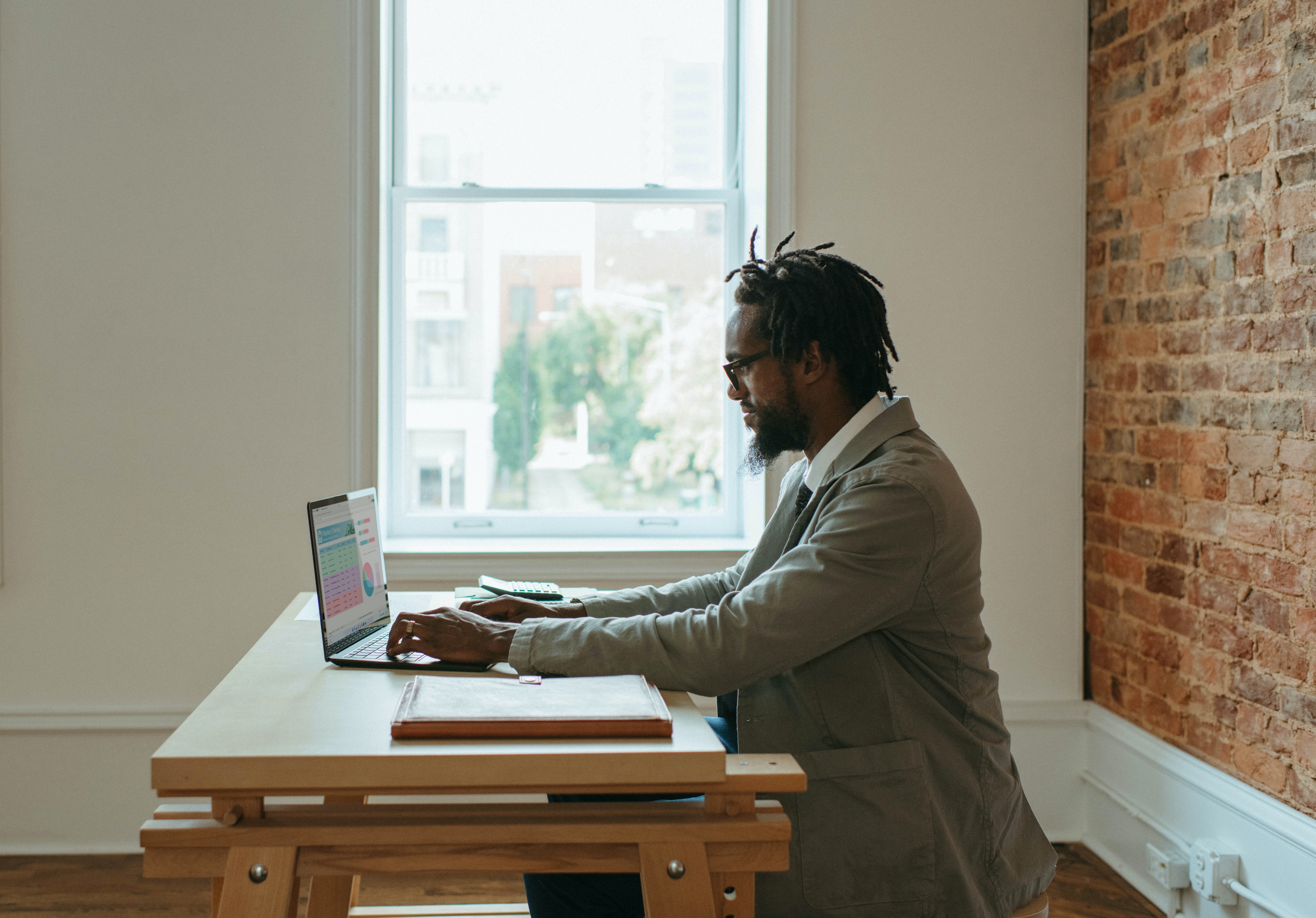 Man sitting at a desk with a laptop in his home office with a window and brick wall.