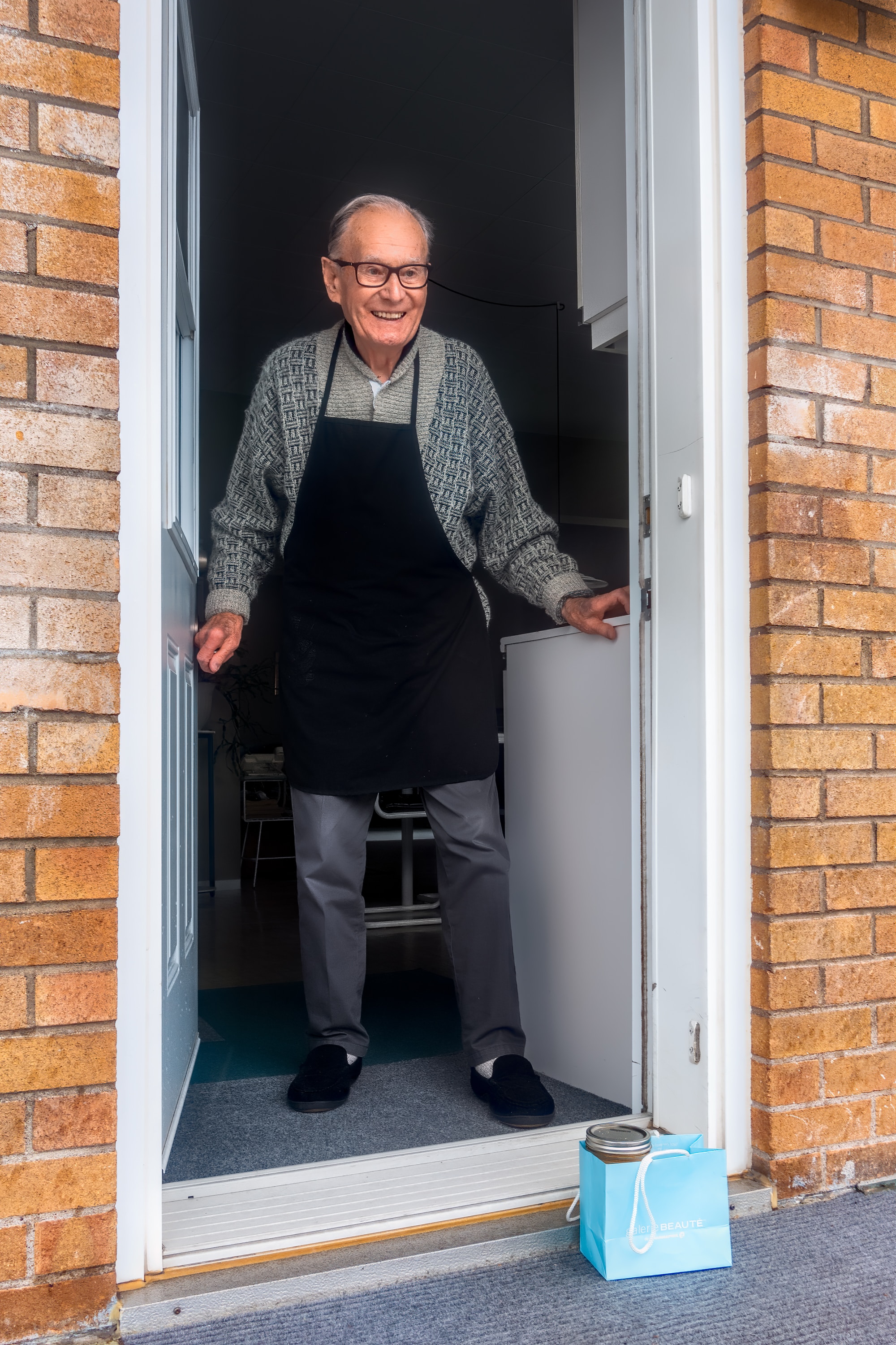 Elderly man wearing an apron standing at his back door.