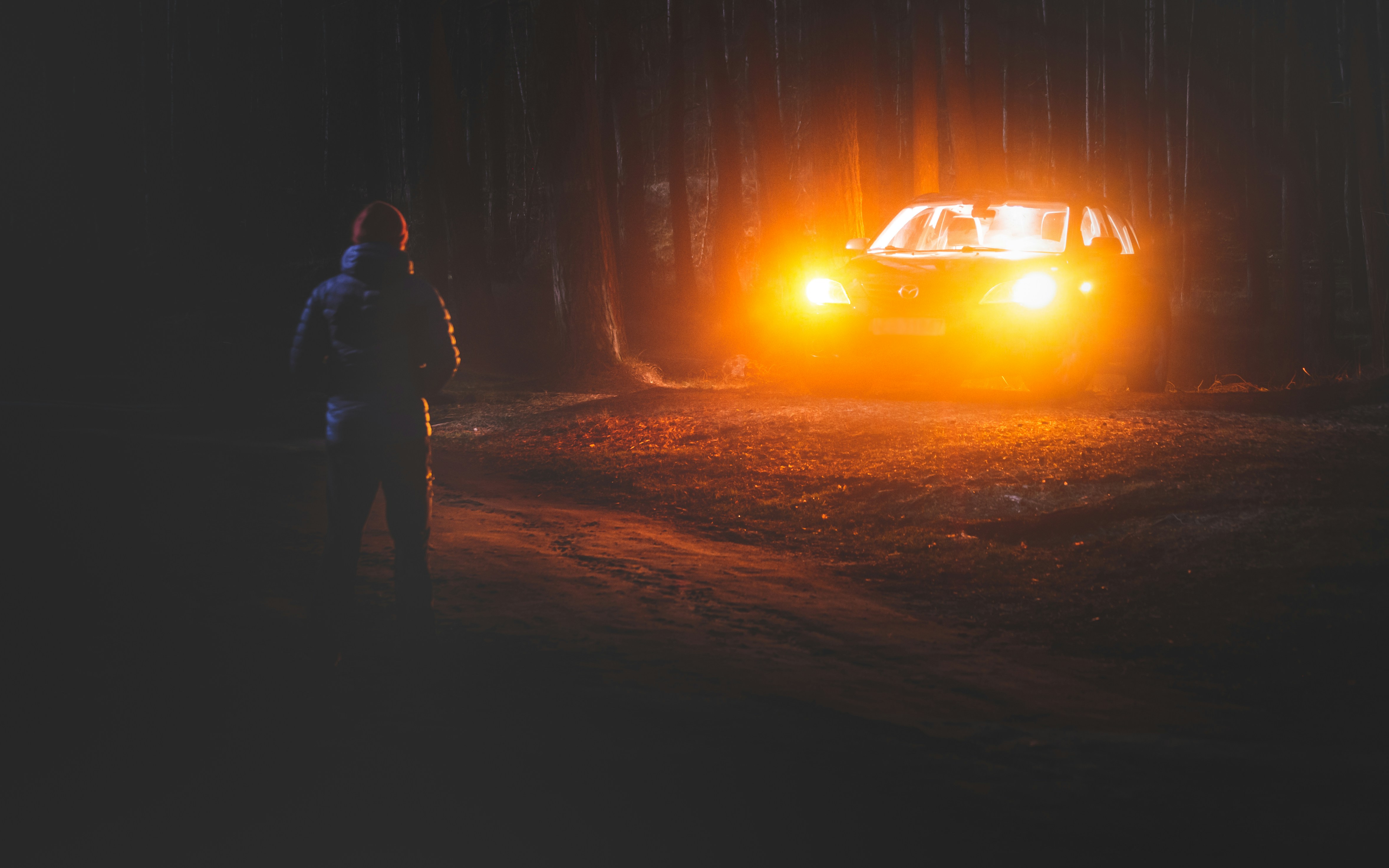 Mysterious man stands in front of a car with headlights on.