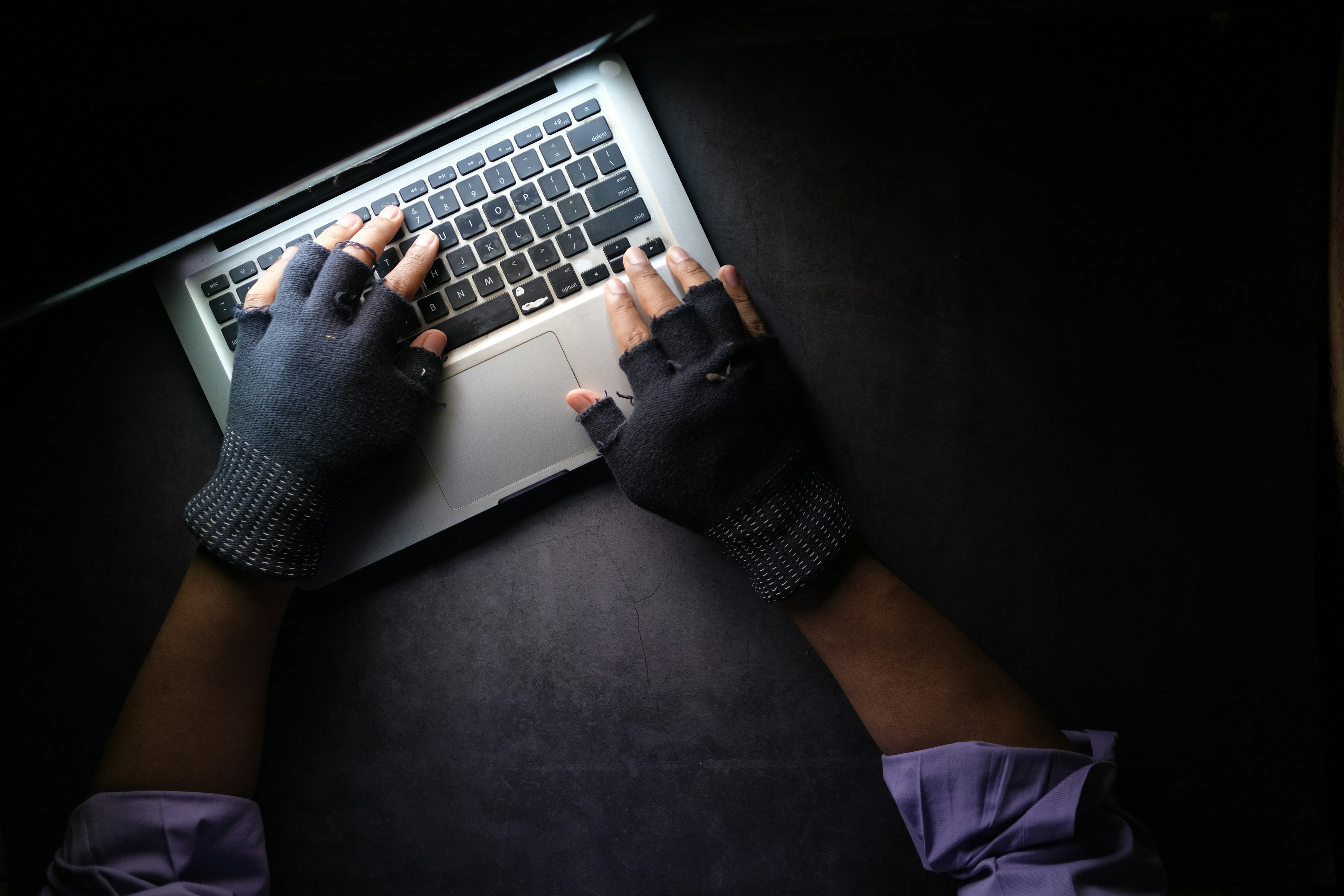 A criminal with fingerless gloves types on a laptop in the dark.