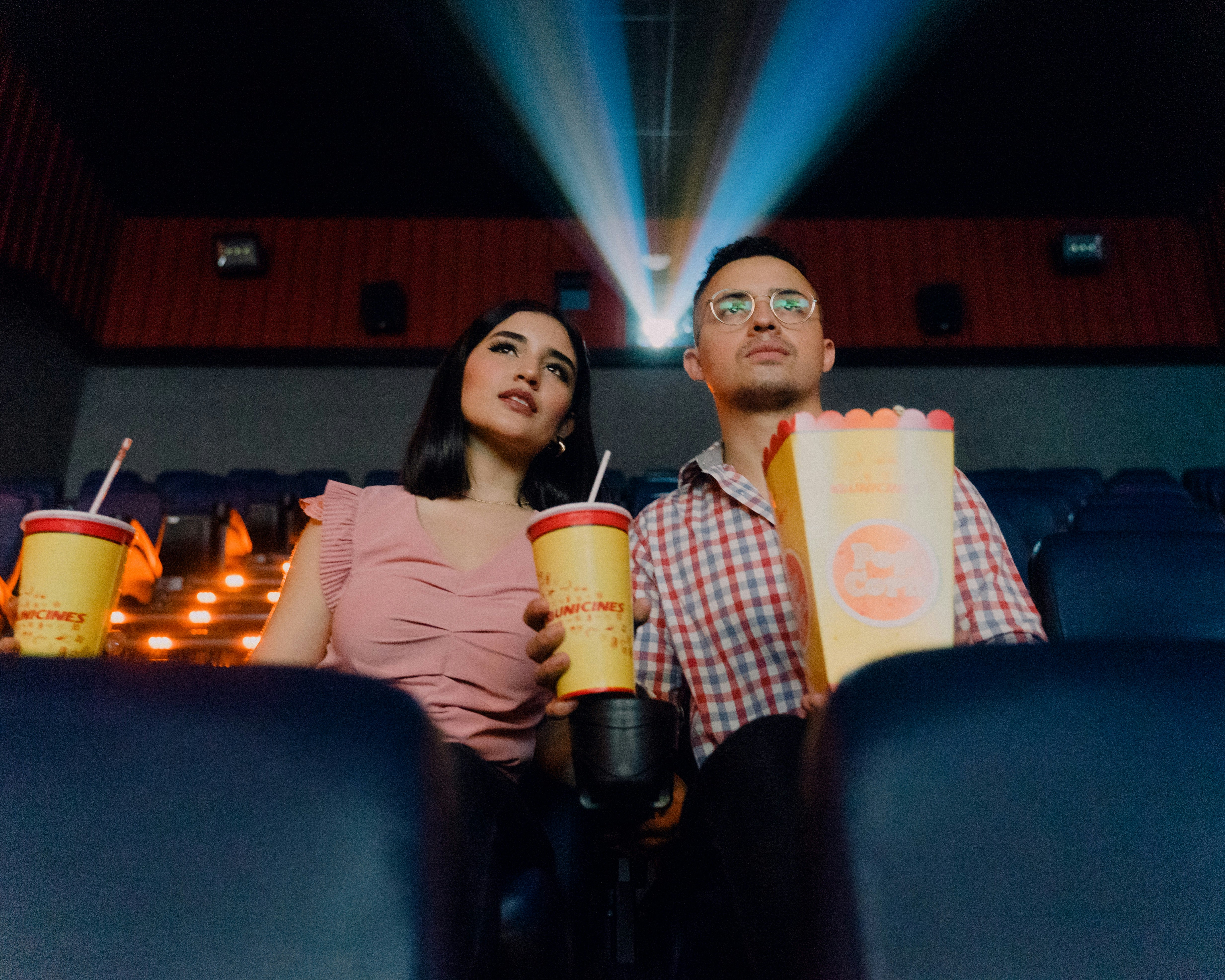 A couple sitting in a movie theatre holding drinks and popcorn.