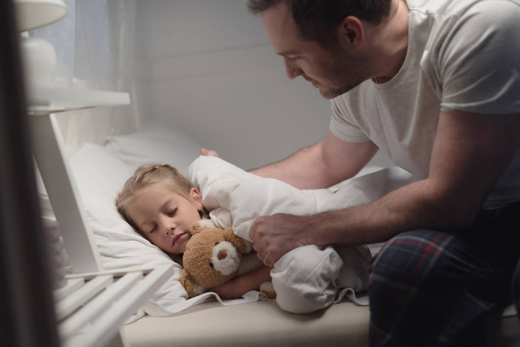 Child sleeping with a stuffed animal as parent tucks them in.