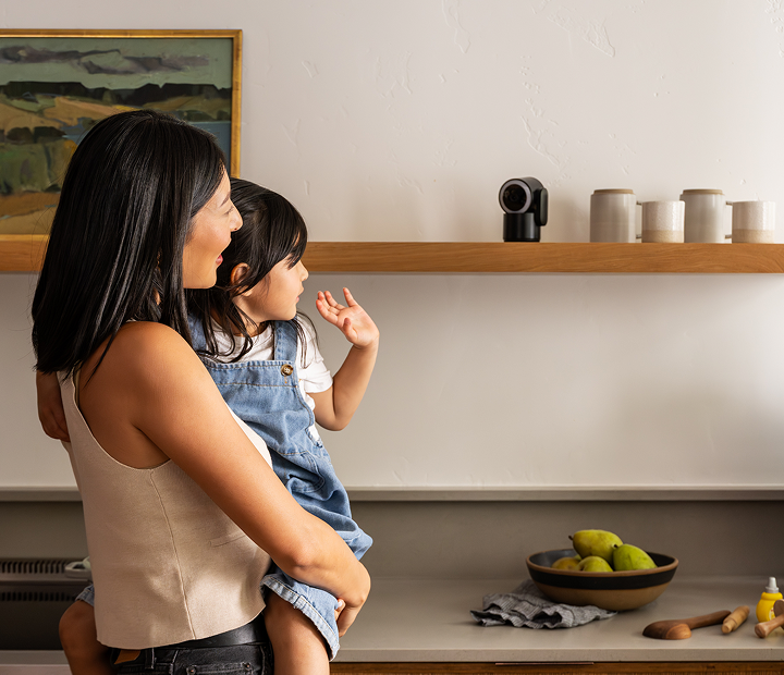 Woman holding child waving to Cove Indoor Cam on shelf