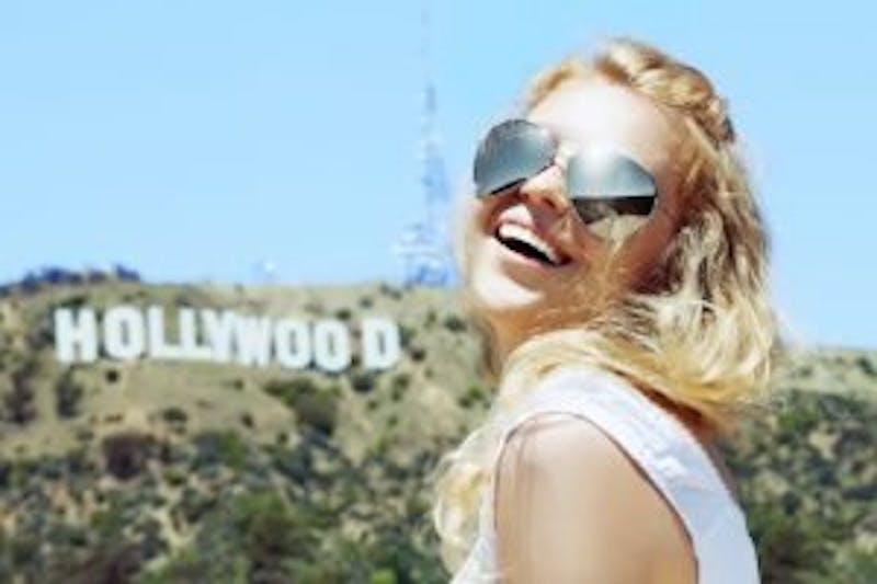 blond woman with sunglasses and a white top smiling in front of a hollywood sign