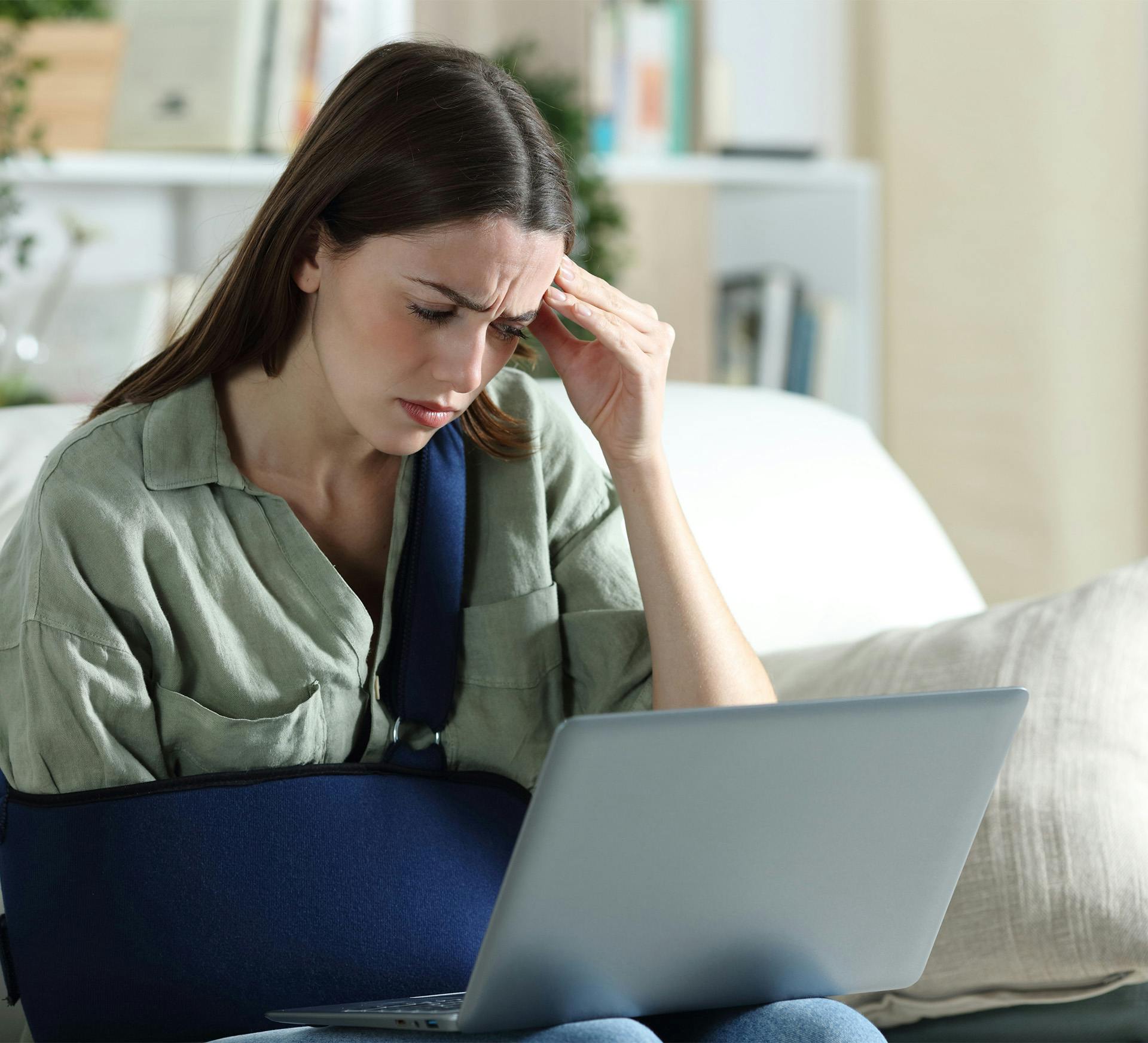 distressed looking woman sitting on a couch with her arm in a sling while looking at a laptop
