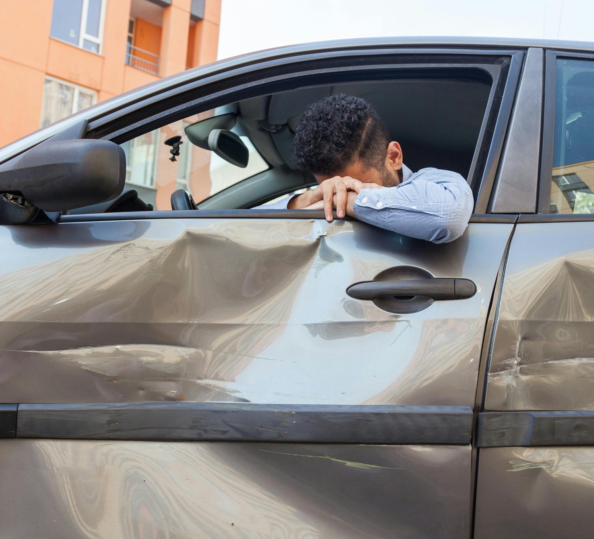 a distressed man sitting in the driver side of damaged vehicle has head resting on top of arms outside of windown