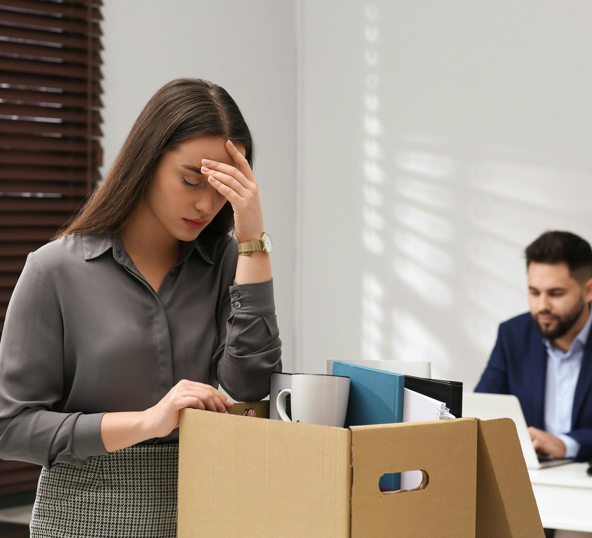 woman in grey shirt looks distressed while packing a box with office supplies from desk