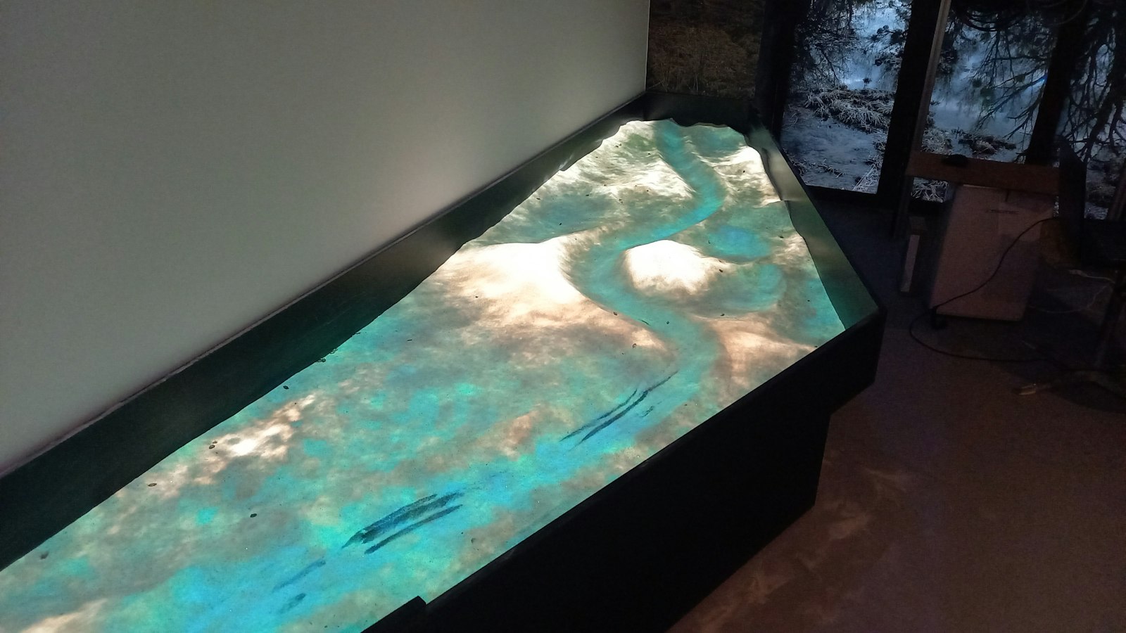 Sandbox during setup, with a pale turquoise projection over a sand river bend in a dark exhibition room.