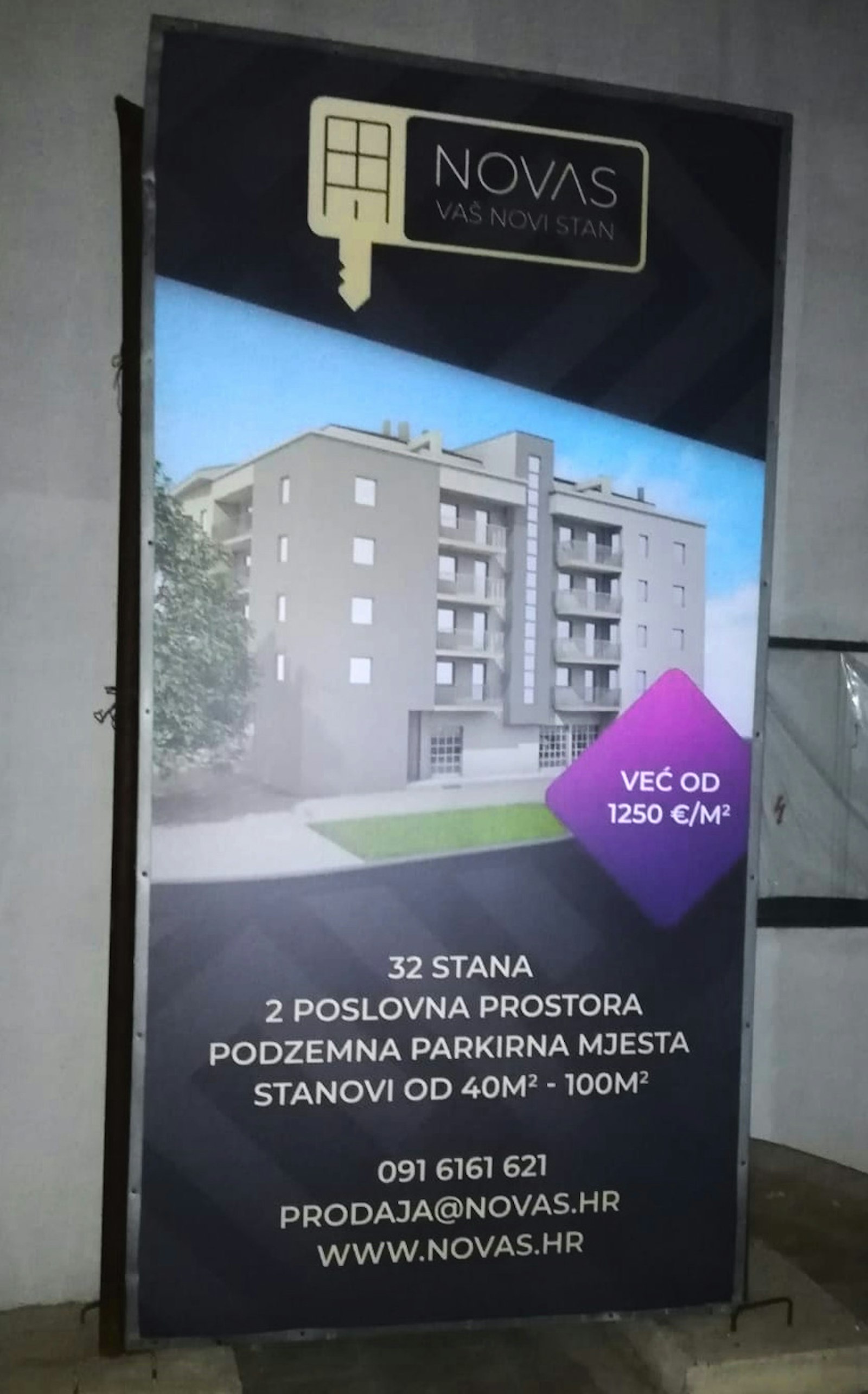Photo of a Novas billboard on site showing the building render, prices from 1250 €/m² and contact details.