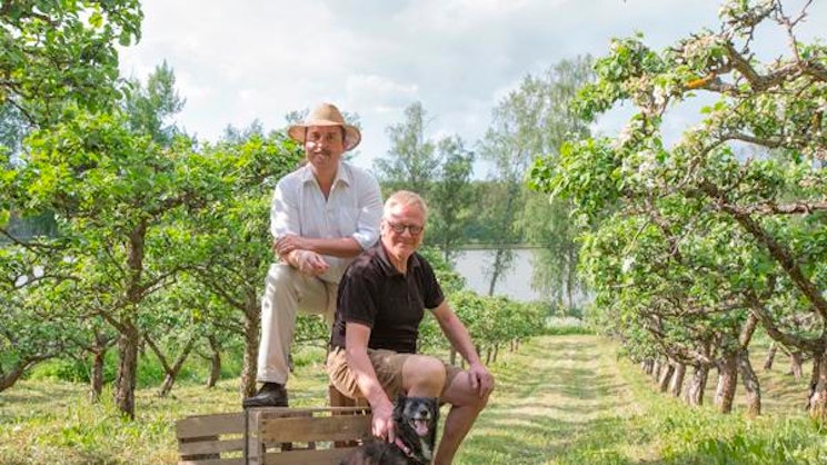 Brinkhall’s-kuohuviinin osakas Juhani Salovaara ja toinen yrityksen edustaja istuvat omenatarhassa koiran kanssa.