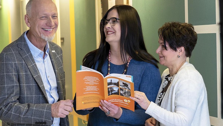 kaksi naista ja mies pitävät kirjaa kädessään