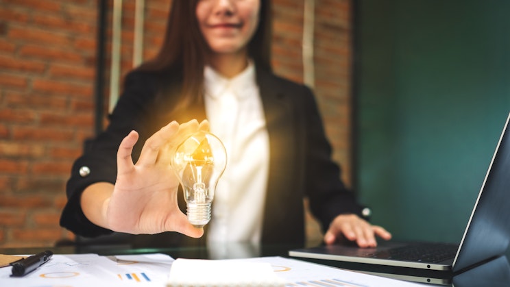 Ihminen pitää hehkulamppua toisella kädellään kannettavan tietokoneen päällä.