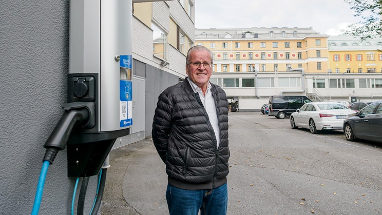 Kai Lahtonen seisoo sähköauton latauspisteen vieressä taloyhtiön pysäköintialueella. Latauslaite näkyy kuvan etualalla rakennuksen seinään asennettuna.