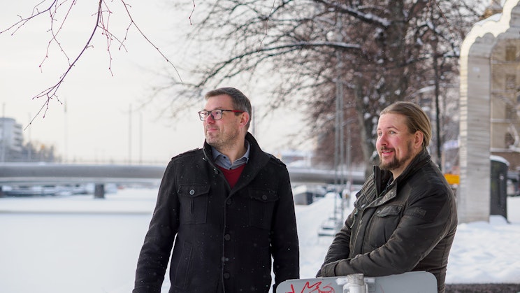 Kuvassa kaksi henkilöä seisoo Aurajoenrannassa.