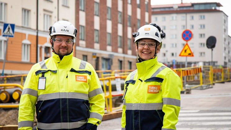 Kuvassa kaksi henkilöä hymyilee kameralle työmaavaatetuksessa