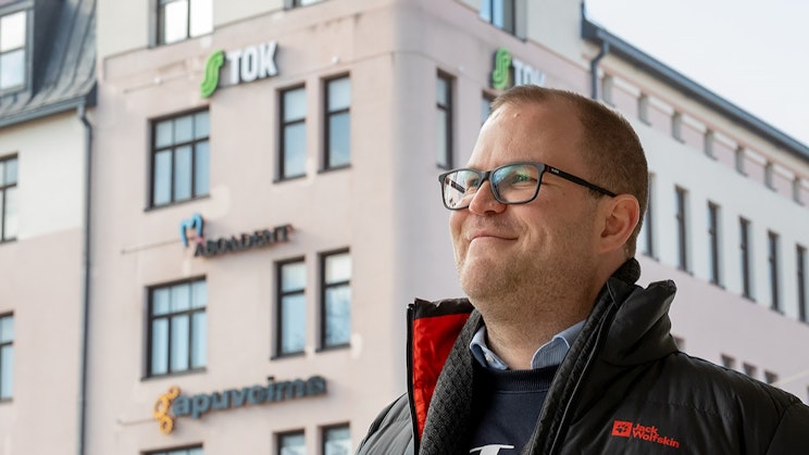 Kuvassa mies hymyilee ja hänen takanaan on TOKin toimistorakennus