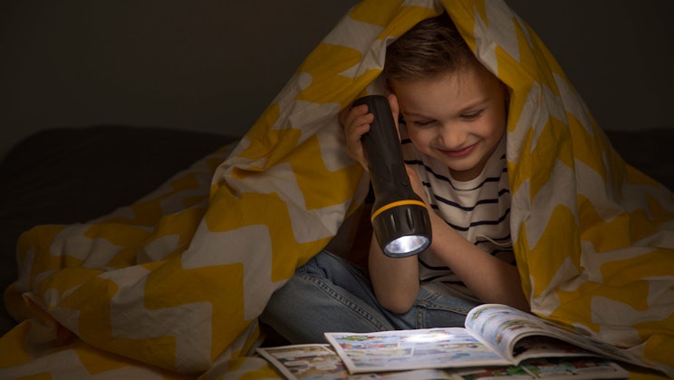Poika lukee lehteä peiton alla taskulampun valossa