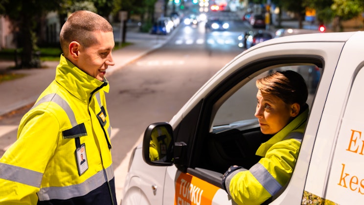 Kuvassa kaksi henkilöä, joista toinen istuu autossa
