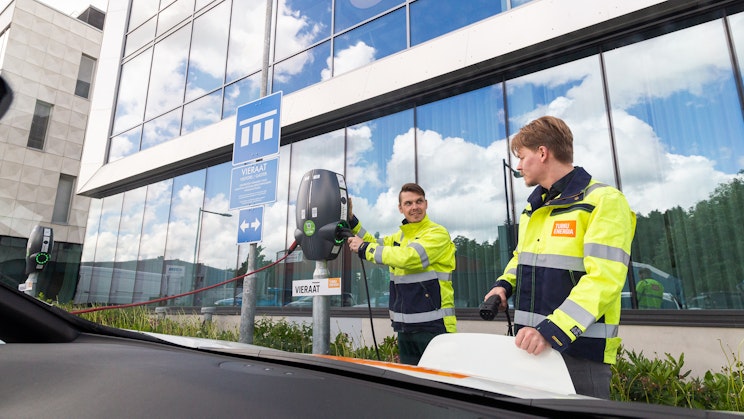 Kuvassa kaksi henkilöä lataavat sähköautoa rakennuksen edessä