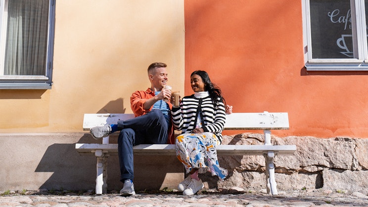 pariskunta juomassa kahvia valkoisella penkillä rapattujen ja värikkäiden talojen edessä