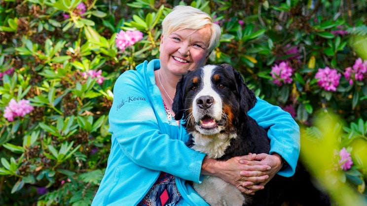 Maarit Haapasaari istuu kukkapensaan edessä ja halaa koiraansa.