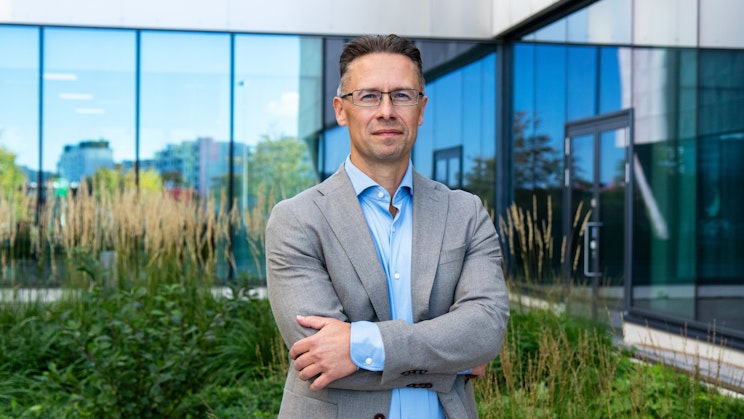 Mies oletettu seisoo kädet puuskassa rakennuksen ja viheriön edessä