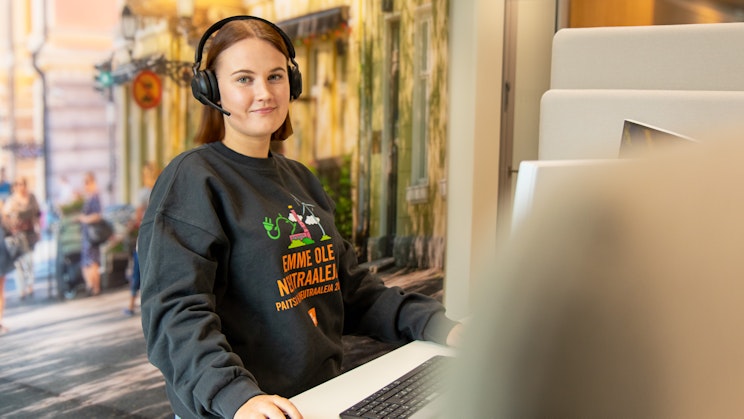 Naisoletettu seisoo tietokoneen ääressä kuulokkeet päässä ja katsoo kameraan