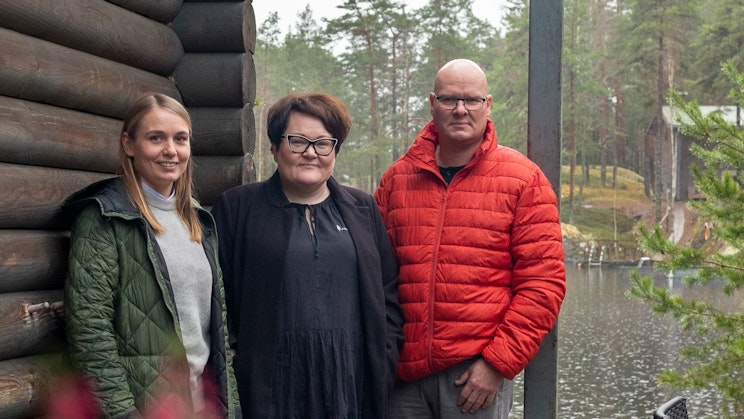 Kolme henkilöä poseeraa kameralle mökin ja rannan edustalla