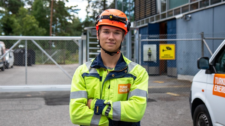 Henkilö, jolla on työvaatteet seisoo kädet puuskassa ja hymyilee kameraan