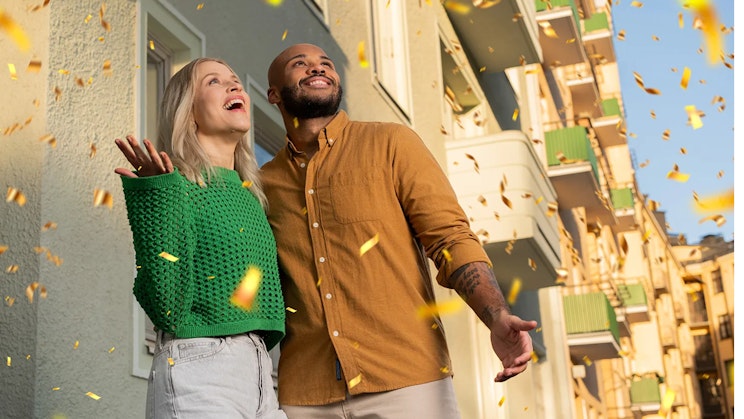 Kaksi henkilöä seisovat kerrostalon edessä konfettisateessa