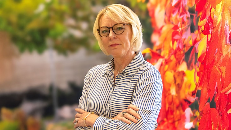 Anne-Mari Niemelä seisoo kädet ristissä raidallisessa paidassa värikkään, punasävyisen syyslehtitaustan edessä.