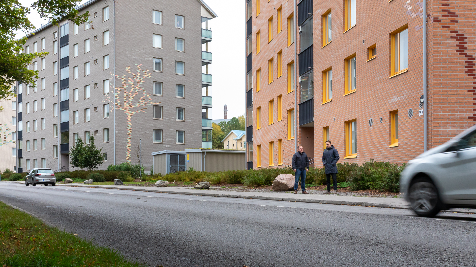 Valokuva Turun Mäntymäessä sijaitsevan Kunnallissairaalantie 36–42 kerrostalojen edustalta. Kuvassa näkyy kaksi rakennusta, jalkakäytävällä seisoo kaksi henkilöä. Harmaan talon julkisivussa on koristeena puun kuvio. Etualalla kulkee asfalttitie, jonka reunassa on liikkeessä auto.