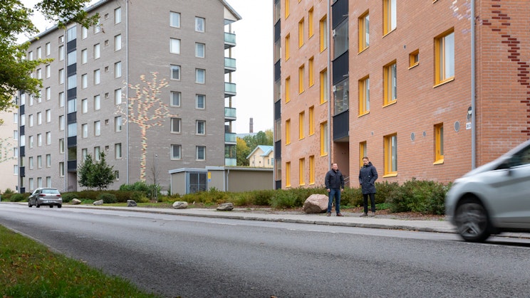 Valokuva Turun Mäntymäessä sijaitsevan Kunnallissairaalantie 36–42 kerrostalojen edustalta. Kuvassa näkyy kaksi rakennusta, jalkakäytävällä seisoo kaksi henkilöä. Harmaan talon julkisivussa on koristeena puun kuvio. Etualalla kulkee asfalttitie, jonka reunassa on liikkeessä auto.