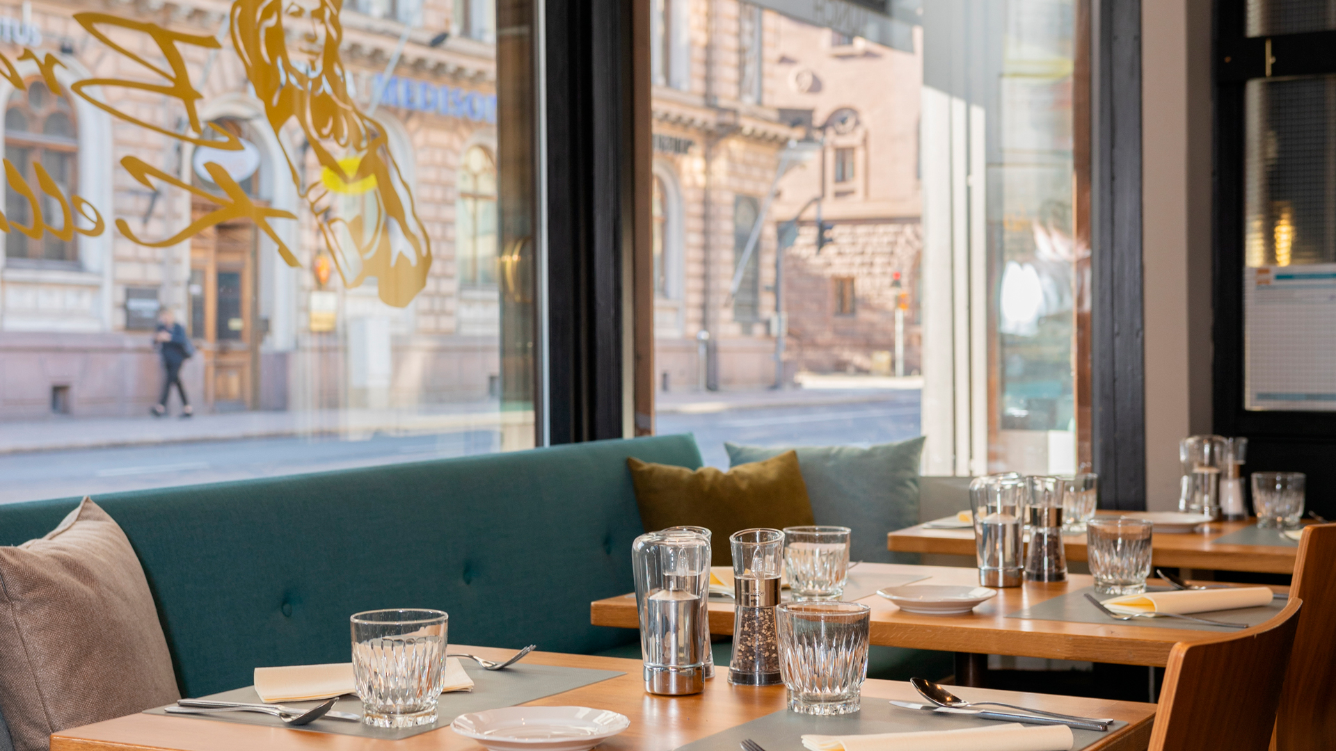 Sisäkuva Turun Brahen Kellarin ravintolasta; etualalla puupöytiä katettuna vesilaseilla, aterimilla ja mausteilla, taustalla turkoosi sohva tyynyineen. Suuren ikkunan läpi näkyy Linnankadun kivirakennuksia ja ohikulkija kadulla kirkkaassa päivänvalossa.