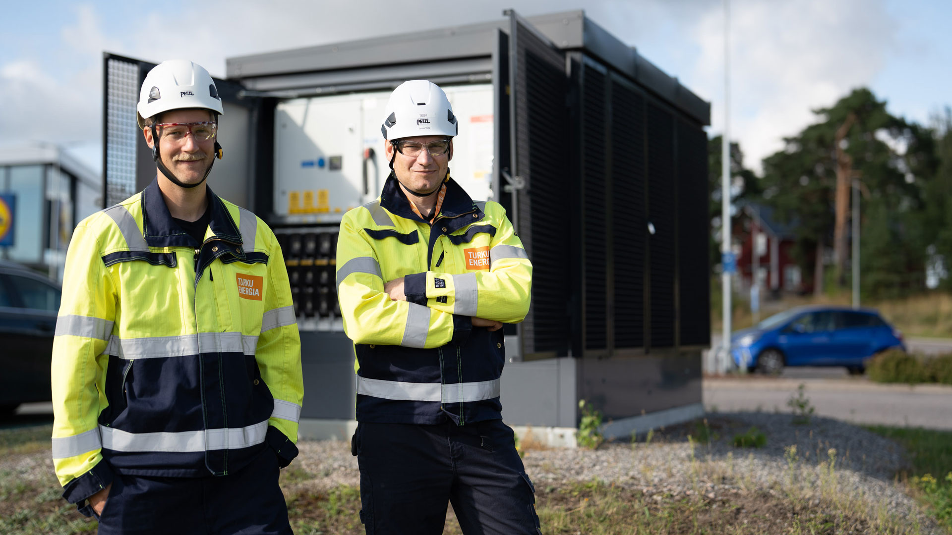 Kaksi Turku Energia Sähköverkkojen työntekijää seisoo ulkona sähköaseman laitteiston edessä, yllään huomiotakit ja valkoiset suojakypärät.