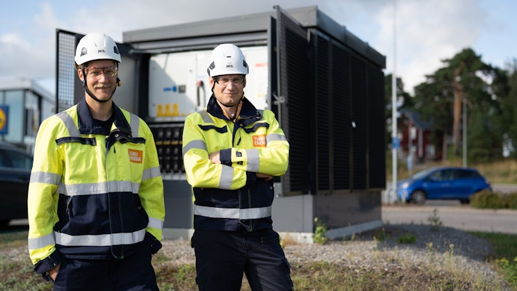 Kaksi Turku Energia Sähköverkkojen työntekijää seisoo ulkona sähköaseman laitteiston edessä, yllään huomiotakit ja valkoiset suojakypärät.