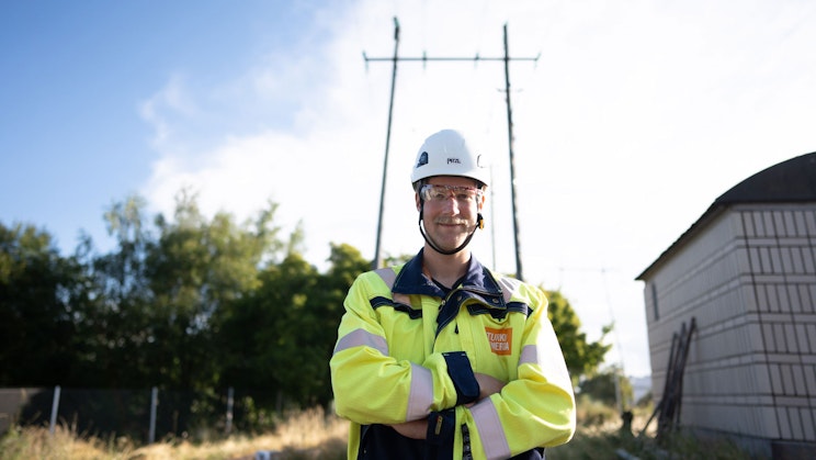 Turku Energia Sähköverkkojen työntekijä seisoo ulkona sähkölinjojen alla ja sähkömuuntamon vieressä..