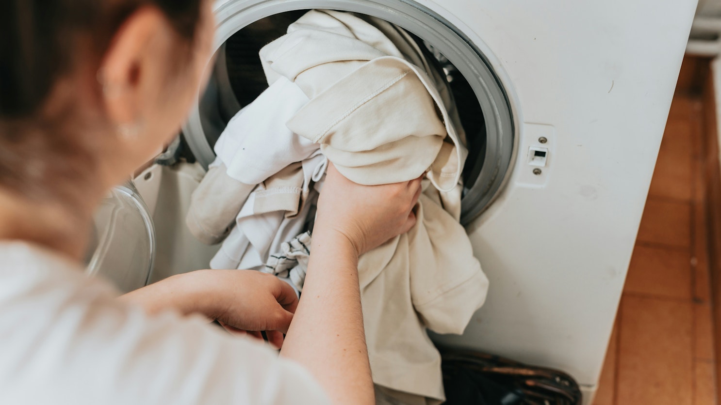 Selän takaa otettu lähikuva, jossa henkilö täyttää edestä täytettävää pyykinpesukonetta vaaleilla vaatteilla.