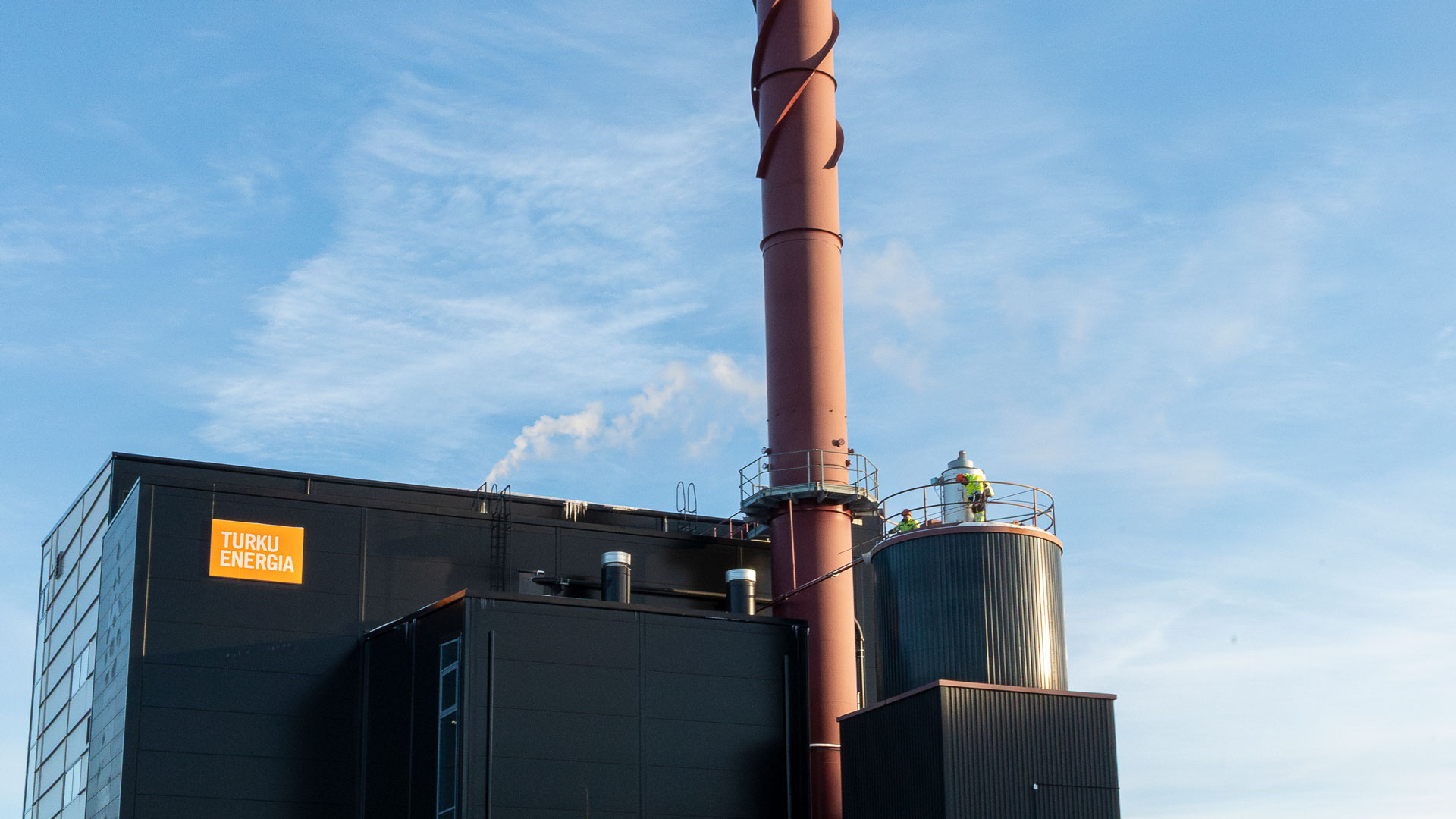 Valokuva Turku Energian tummajulkisivuisesta Artukaisten höyryntuotantolaitoksesta ja sen korkeasta punaruskeasta savupiipusta kirkkaan taivaan alla. Rakennuksen katolla näkyy kaksi suojavarusteisiin pukeutunutta työntekijää.