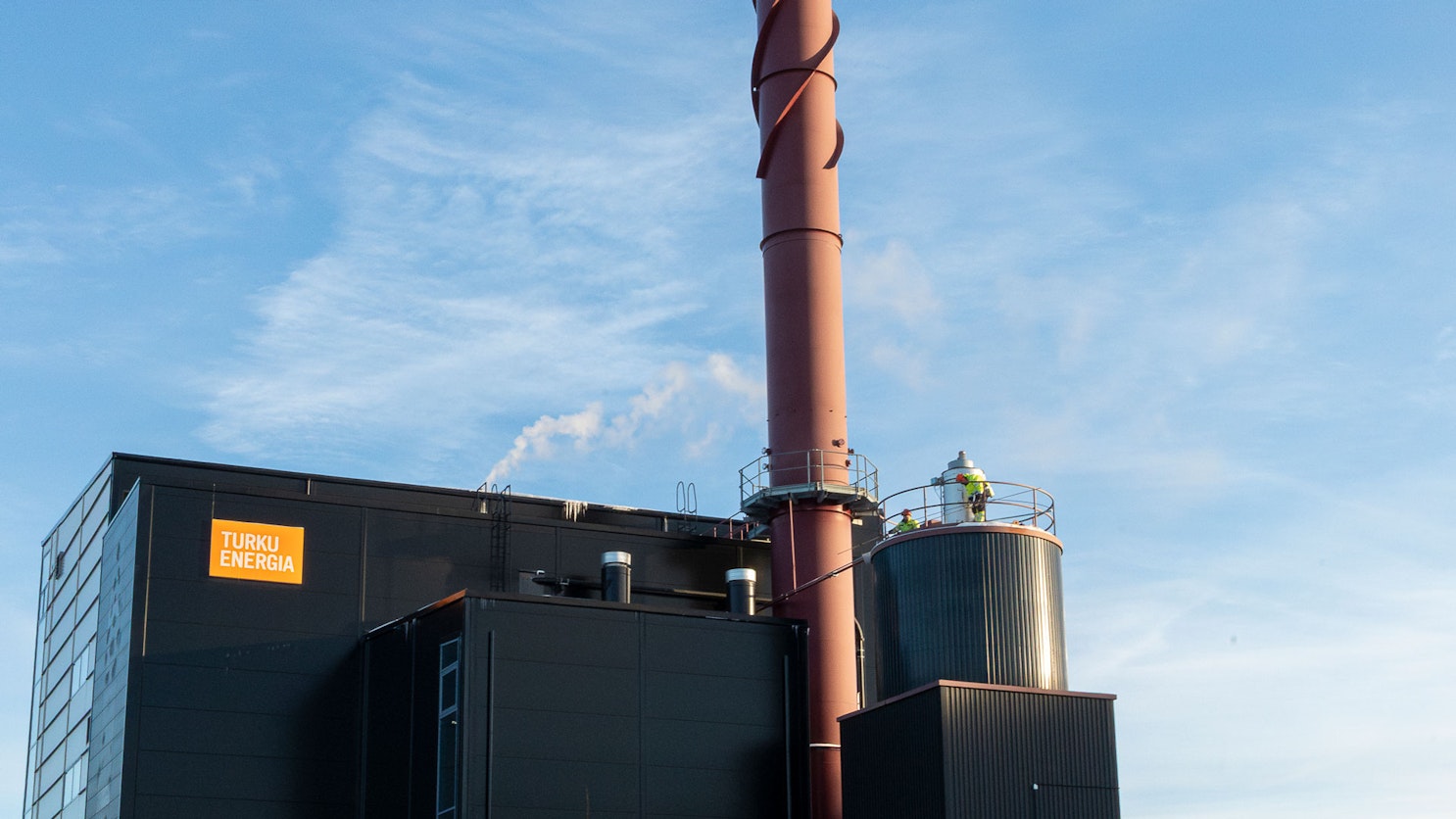 Valokuva Turku Energian tummajulkisivuisesta Artukaisten höyryntuotantolaitoksesta ja sen korkeasta punaruskeasta savupiipusta kirkkaan taivaan alla. Rakennuksen katolla näkyy kaksi suojavarusteisiin pukeutunutta työntekijää.