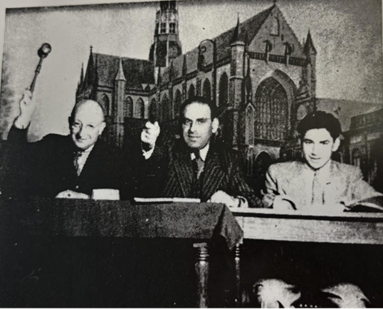 Three men seated at a table during an auction or meeting, with one man holding a gavel aloft. Behind them is a large photograph or backdrop of a Gothic-style cathedral.