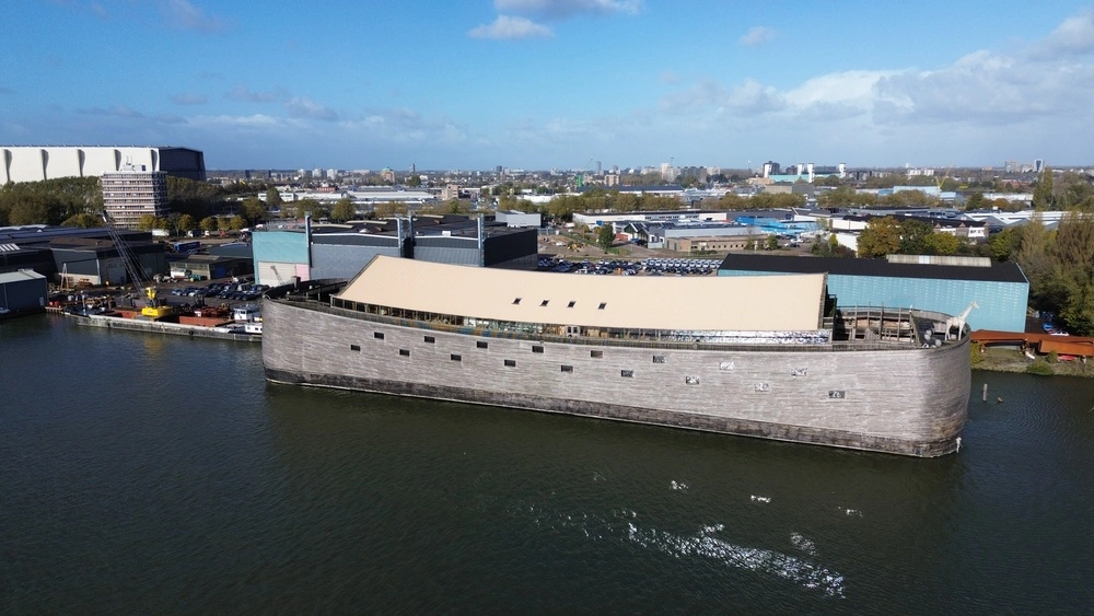 A large wooden replica of Noah’s Ark floating on a river, with industrial buildings and a cityscape visible in the background under a partly cloudy sky.