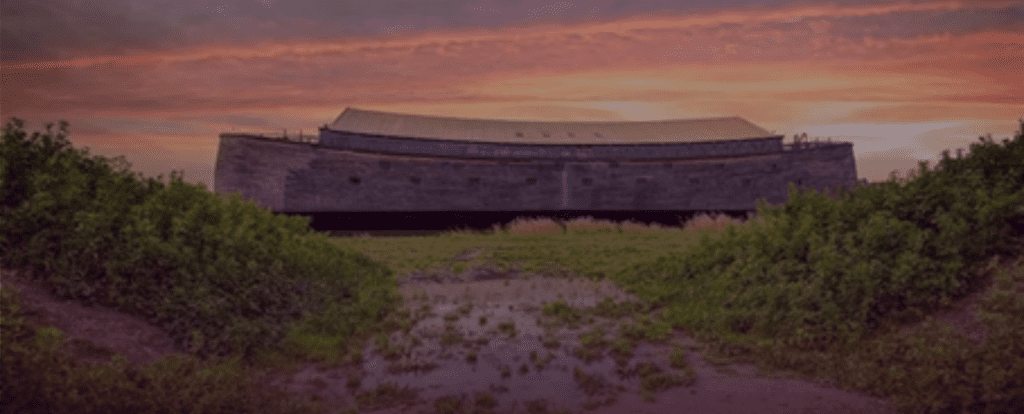 A large wooden ark structure resting on land, surrounded by grass and puddles, with a dramatic sunset sky in the background.