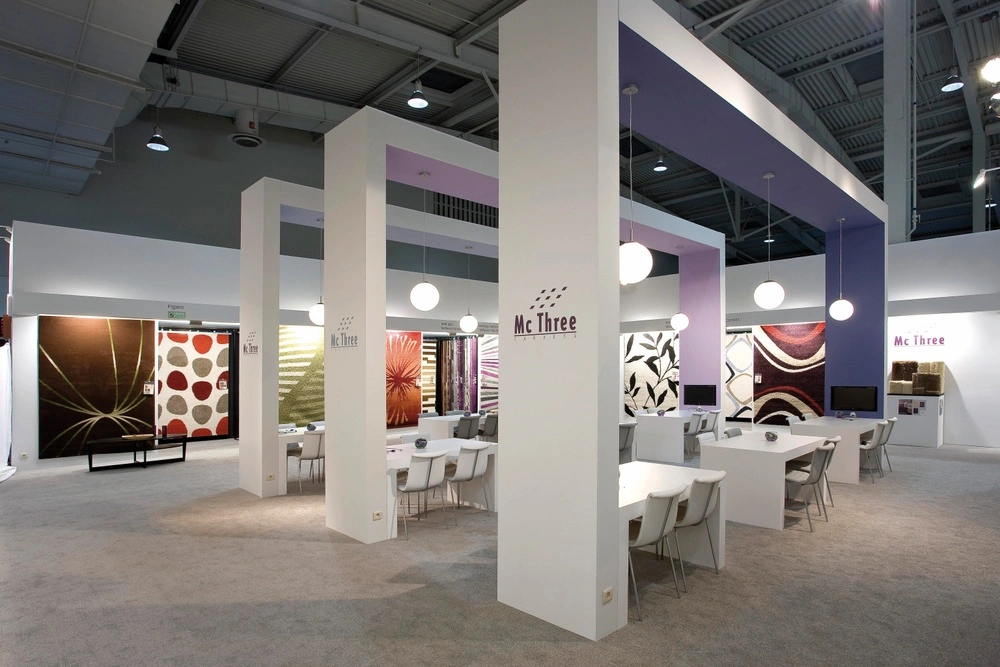 Modern exhibition booth for Mc Three, featuring large white pillars, hanging lights, tables with chairs, and colorful patterned rugs displayed on the walls
