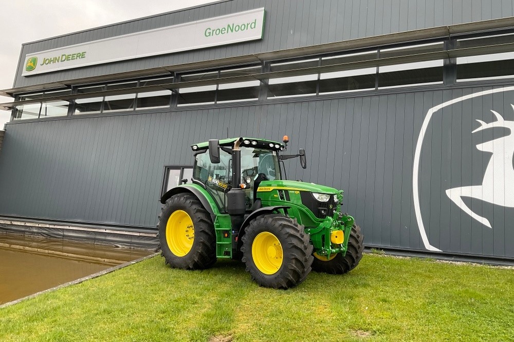 A green and yellow John Deere tractor parked on a grassy area in front of a GroeNoord building with John Deere signage and logo on the wall