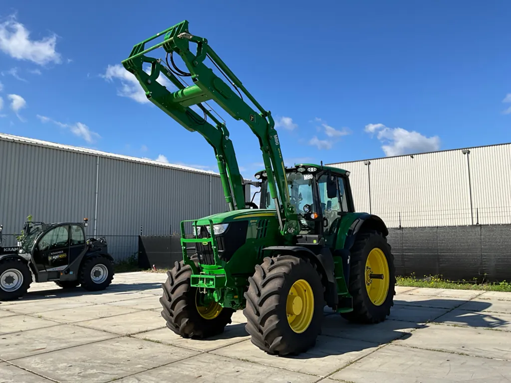 A green John Deere tractor equipped with a front loader attachment, parked on a concrete surface near an industrial building under a clear blue sky.
