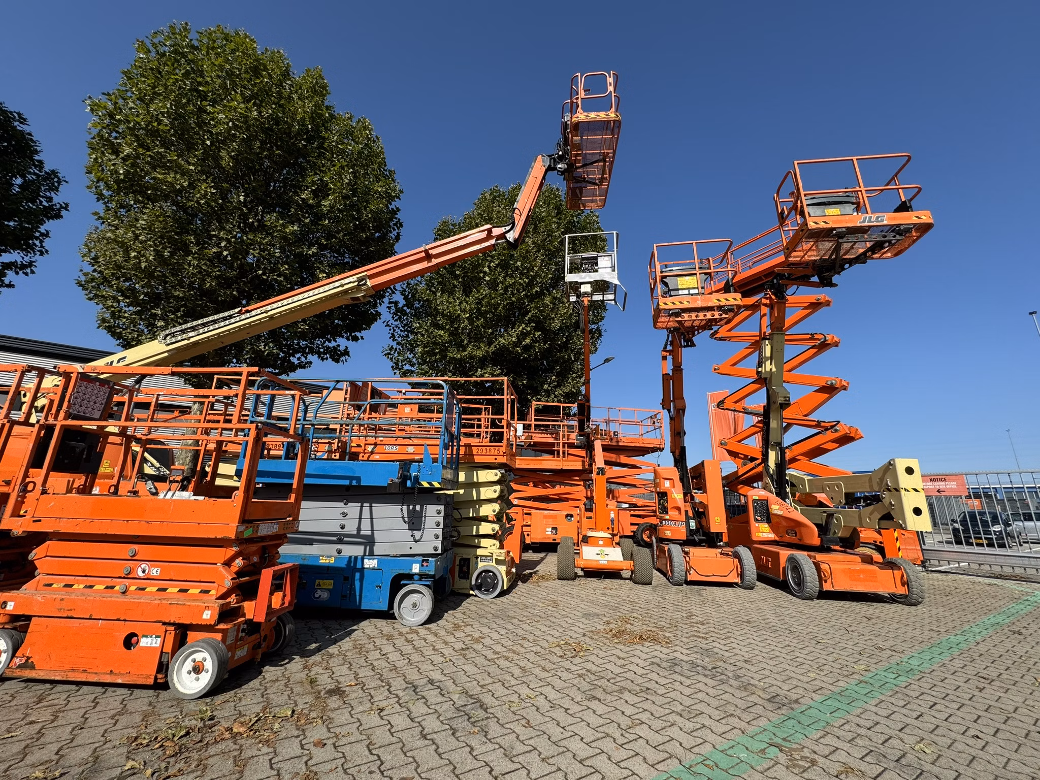 Several orange and blue aerial work platforms and scissor lifts parked outdoors on a paved lot, with some lifts extended under a clear blue sky.